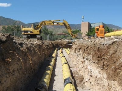Excavator and heavy equipment at a construction site with yellow pipes laid in a long trench under clear blue sky.