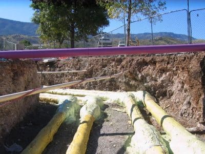 Exposed underground pipes marked with yellow and purple insulation in a trench at a construction site, with fencing and mountains visible in the background.