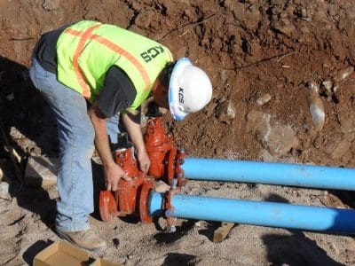 A construction worker in a safety vest and helmet adjusts valves on two blue water pipes at an outdoor work site.