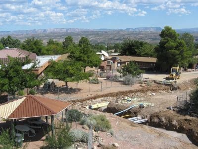 A construction site with earthmoving equipment near buildings, trees, and a covered picnic area, with mountains visible in the background under a partly cloudy sky.